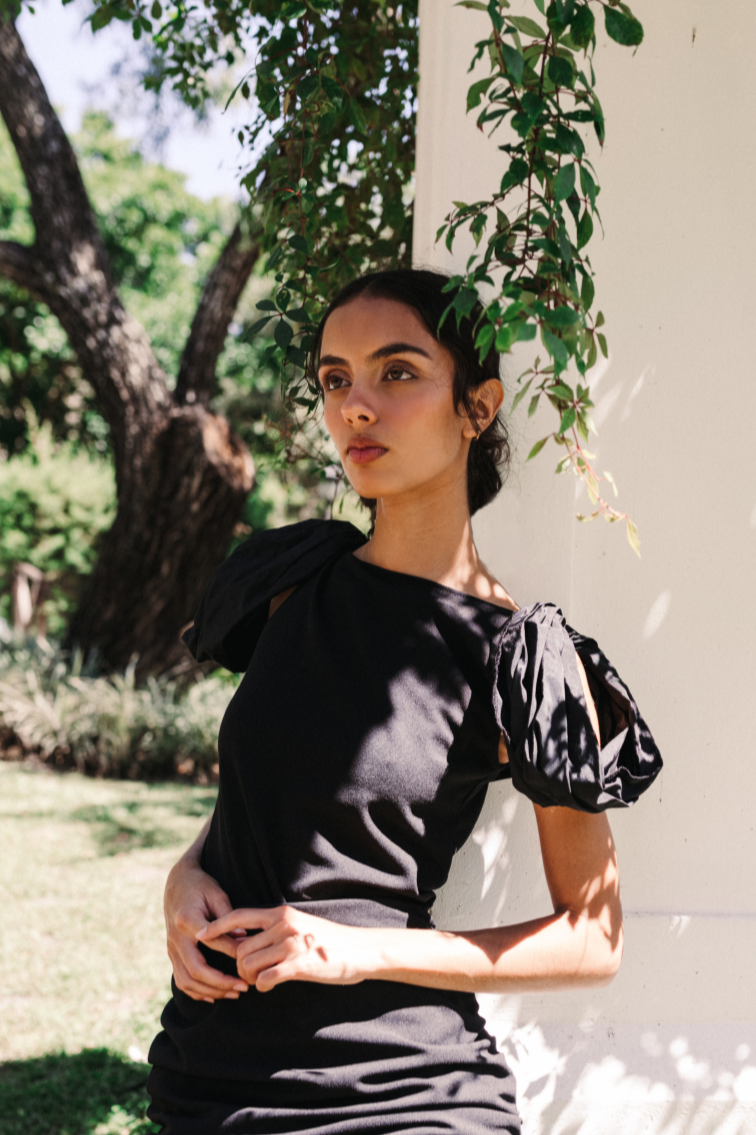 Model wearing a black handmade dress with puffed sleeves by Monica Arguedas, posing against a white column surrounded by greenery in El Salvador