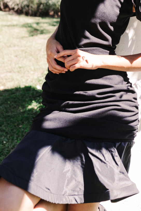 Model wearing a black handmade dress with puffed sleeves by Monica Arguedas, posing against a white column surrounded by greenery in El Salvador