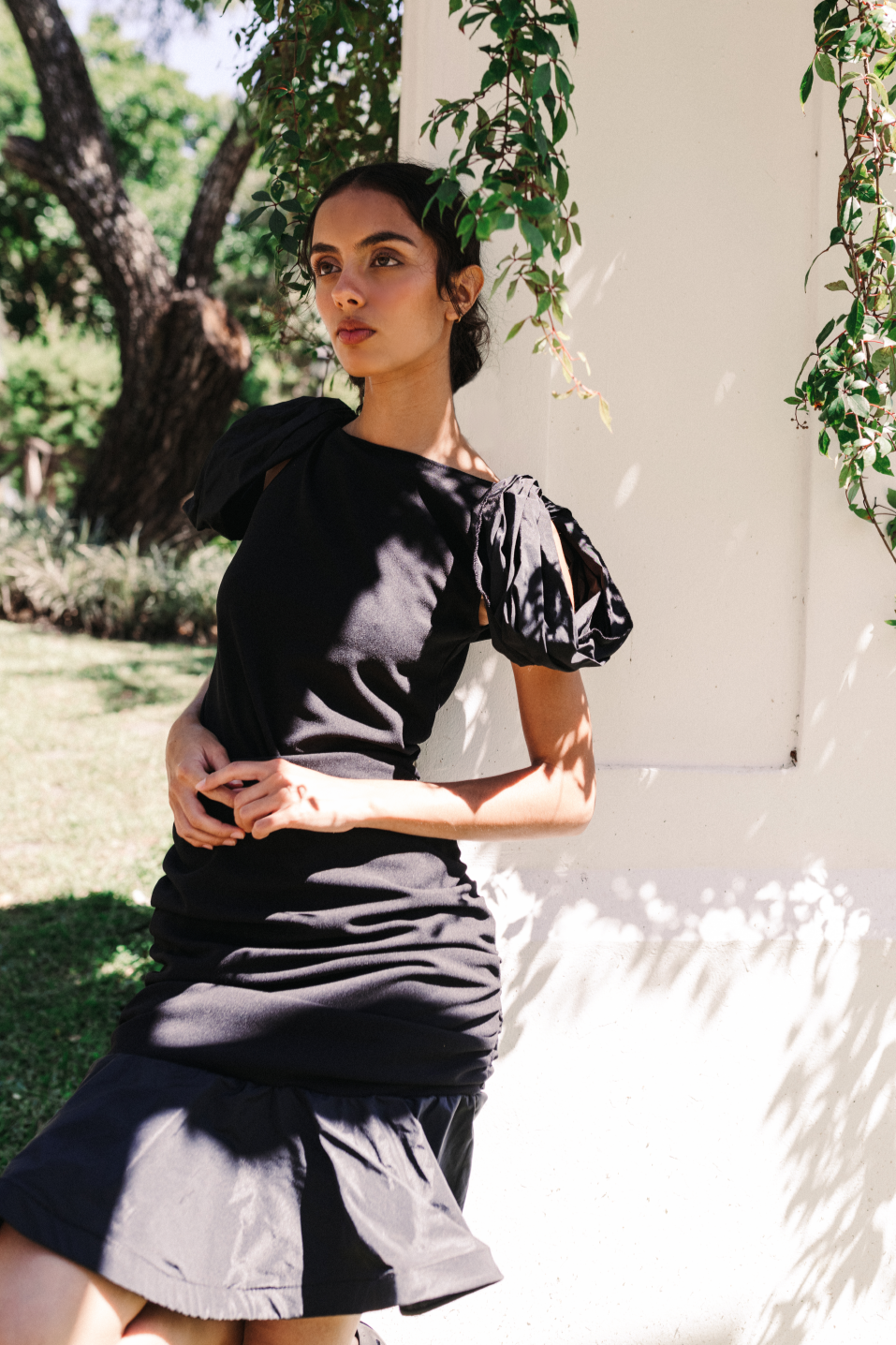 Model wearing a black handmade dress with puffed sleeves by Monica Arguedas, posing against a white column surrounded by greenery in El Salvador