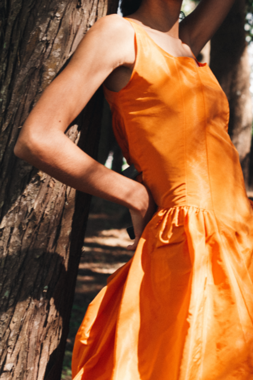 Model wearing a vibrant orange handmade dress by Mónica Arguedas, leaning against a tree in a forest in El Salvador