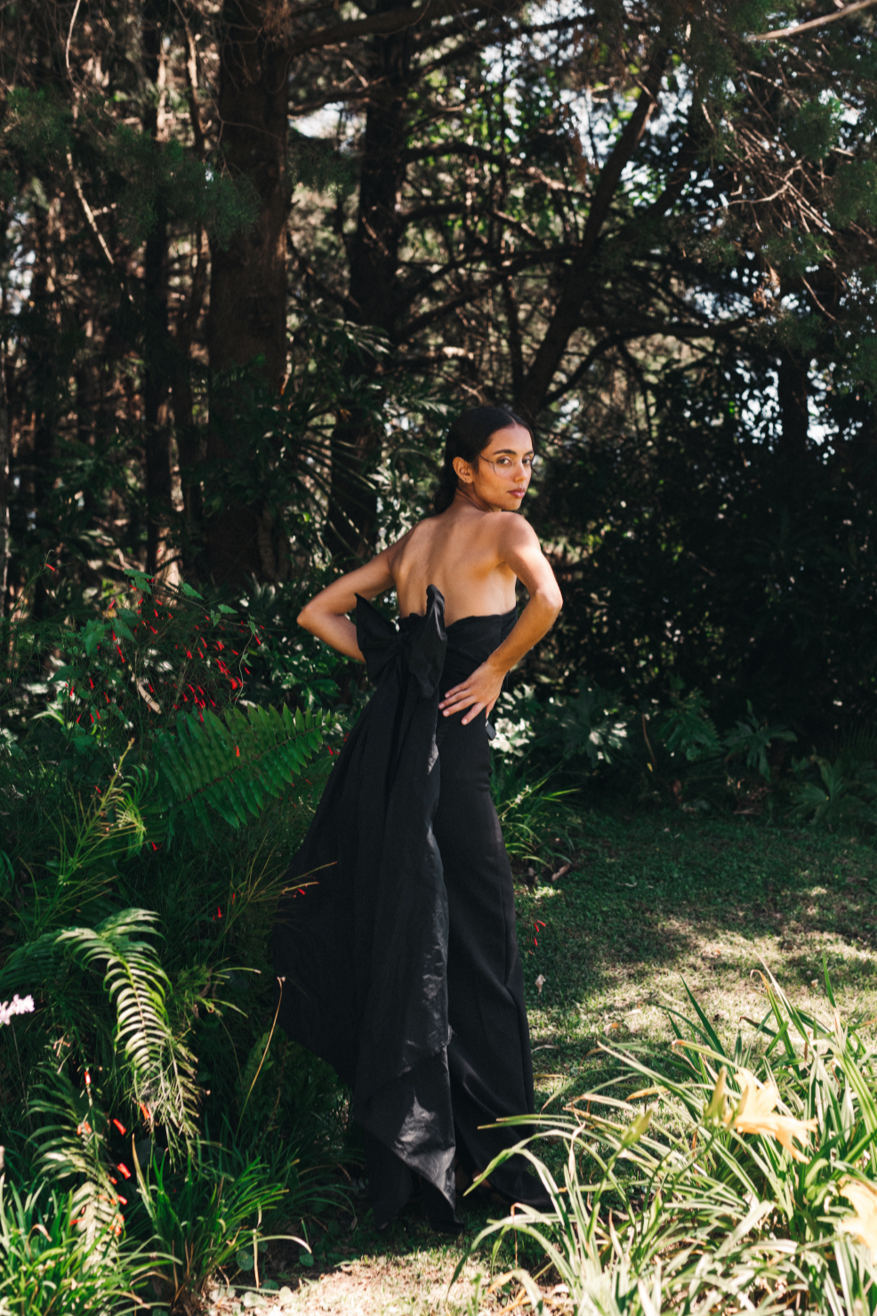 Model wearing a long sleeveless black dress with a big decorative bow on the back surrounded by a salvadoran forest.