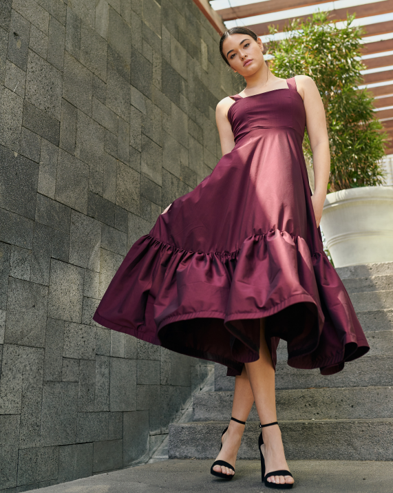 A model wearing a long burgundy dress with a ruffled panel in the bottom by Monica Arguedas.