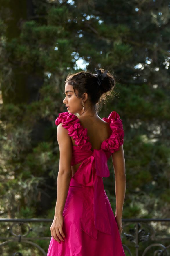 A model wearing a bright pink top with scrunched straps and a bow in the back by Monica Arguedas.