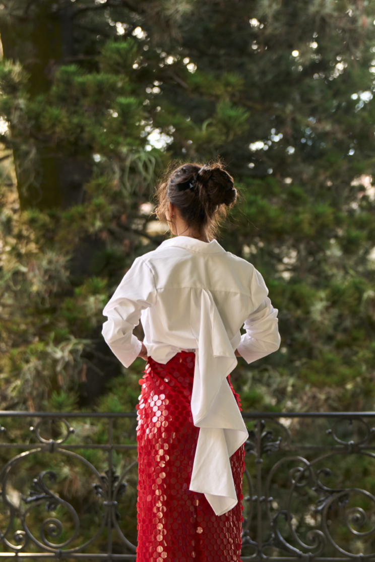 A model wearing a white blouse with a draped panel on the back and slong slevees by Monica Arguedas.