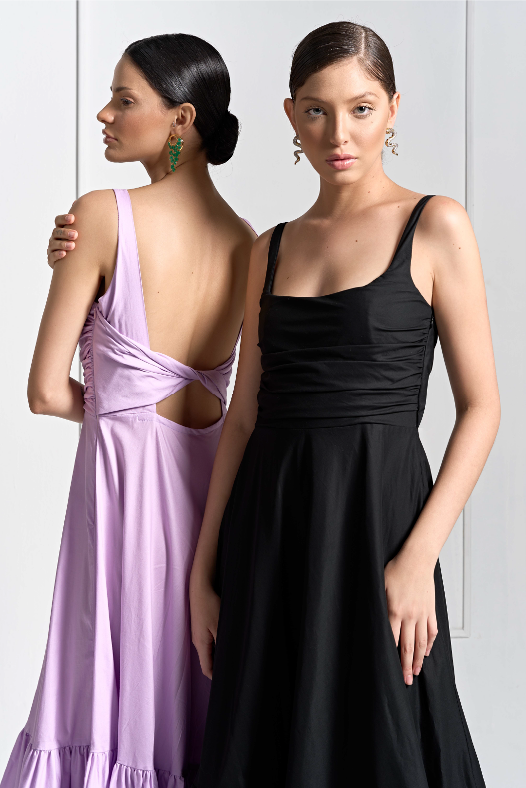 Both models are wearing a dress with ruched detailing at the waist and an open back and twist detail, revealing a subtle cut-out above the waist, one in lilac and the other in black. Both looks made by Monica Arguedas.