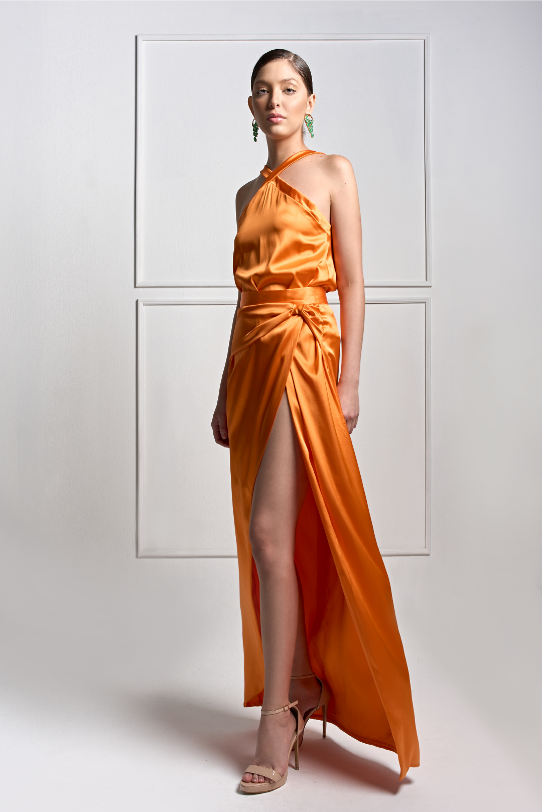 Model wearing a vibrant orange satin set featuring a one-shoulder halter neckline top, draped waist detail and a high front slit skirt by Monica Arguedas.