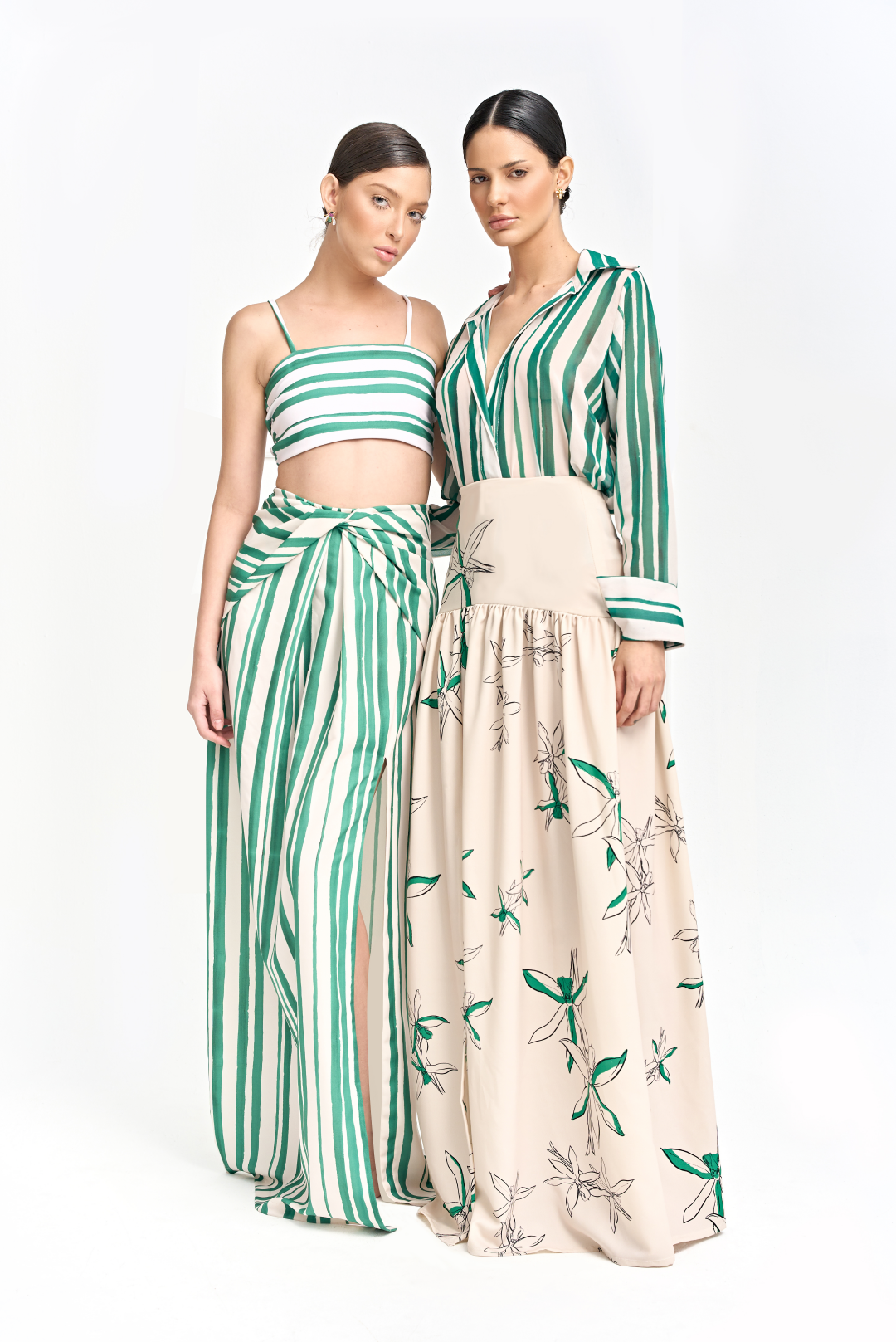 The model on the left wears a green and white striped crop top and matching high-waisted maxi skirt with a side slit. The model on the right wears a green and white striped button-up blouse tucked into a high-waisted beige maxi skirt with green and black floral print. Both looks made by Monica Arguedas.