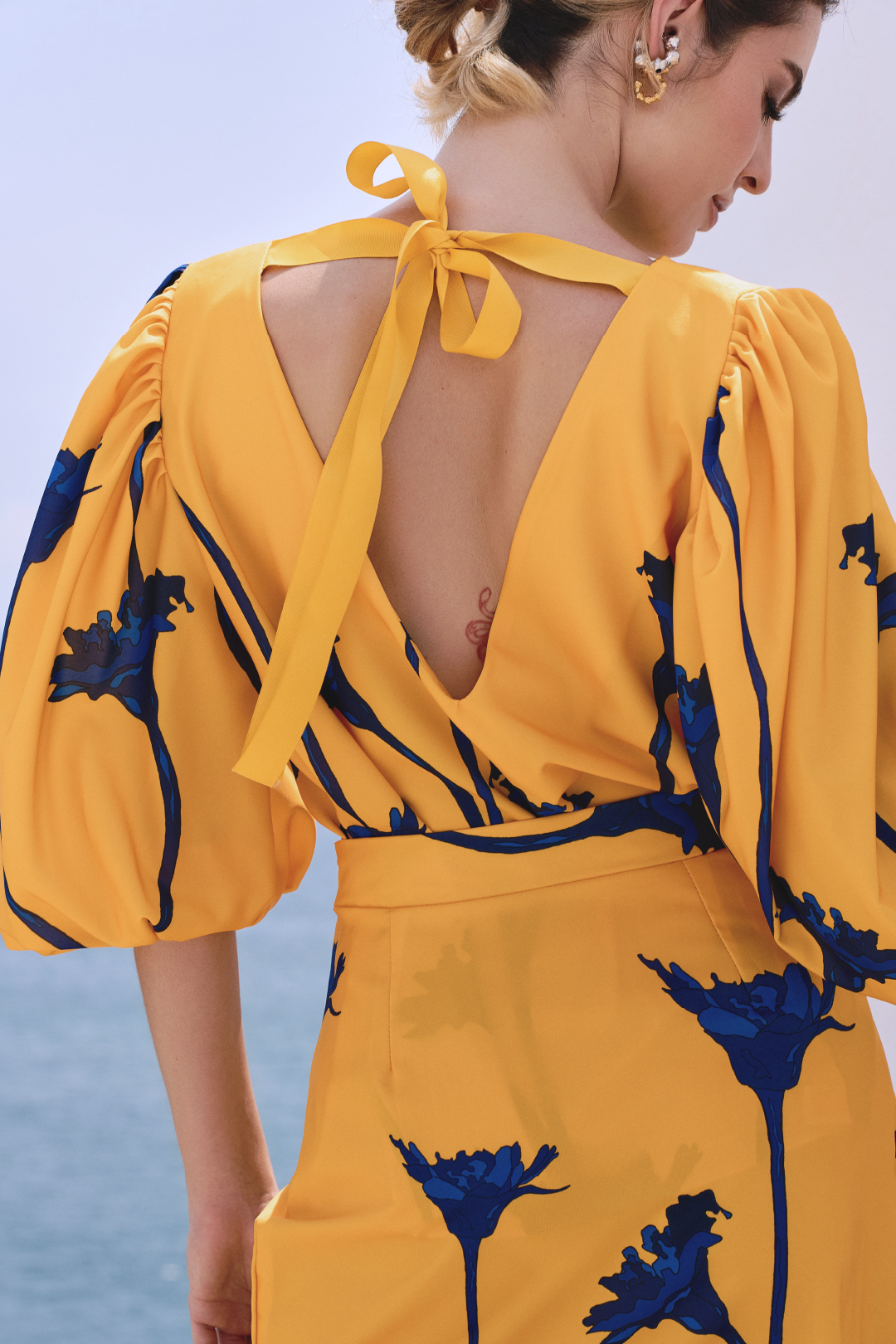 A model wearing a yellow top with blue flowers and a bow detail on the back by Monica Arguedas